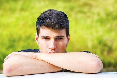 Head and arms shot of handsome attractive young man looking at camera outdoor, leaning on flat surface with head resting on handsの写真素材