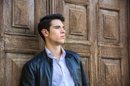 Handsome young man outdoors in front of old house's wood entrance door, looking away to a sideの写真素材
