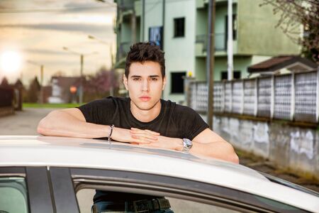 Serious young man sitting on his car's door, resting on the roof and looking at cameraの写真素材