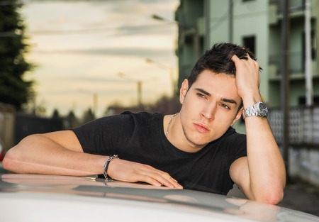 Serious young man sitting on his car's door, resting on the roof and looking away