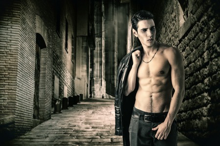 Portrait of a Young Vampire Man in an Open Black Leather Jacket, Showing his Chest and Abs, Looking to Right, on Dark Background.の写真素材