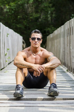 Handsome, serious muscleman sitting against wood fence outdoors, shirtlessの写真素材