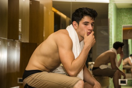 Shirtless handsome muscular male athlete resting on bench in gym&#39;s locker room after workoutの写真素材
