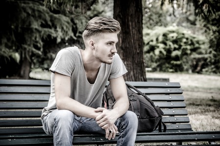 Handsome blond young man sitting on green, wooden park benchの写真素材