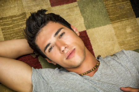 Cool dark-haired handsome young man laying on the floor over colorful rug, looking at camera, hand under his head