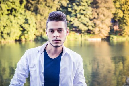 Portrait of contemplative light brown haired young man wearing white shirt beside picturesque river or lake, looking away to a side during fallの写真素材