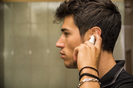 Head and Shoulders Close Up of Attractive Young Man with Dark Hair Spraying Ear Spray into Ears as part of Morning Grooming Routineの写真素材
