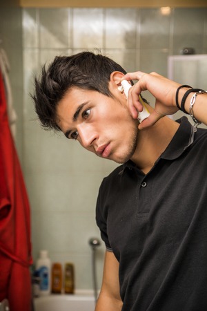 Head and Shoulders Close Up of Attractive Young Man with Dark Hair Spraying Ear Spray into Ears as part of Morning Grooming Routineの写真素材