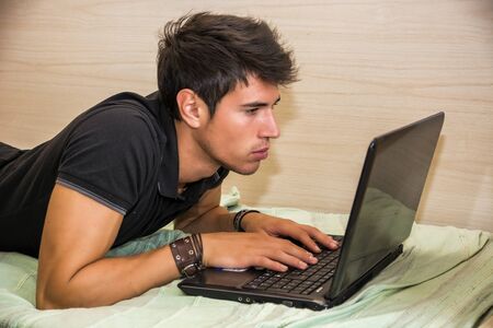 Attractive Young Man with Serious Expression, with Laptop on Bed Working on his Start-up Business - Young Male College or University Student Doing Homework, in Bedroomの写真素材