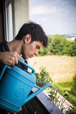 Attractive Young Man on Apartment Balcony Watering Plants in Box from Blue Watering Can on Sunny Day with Field in Backgroundの写真素材