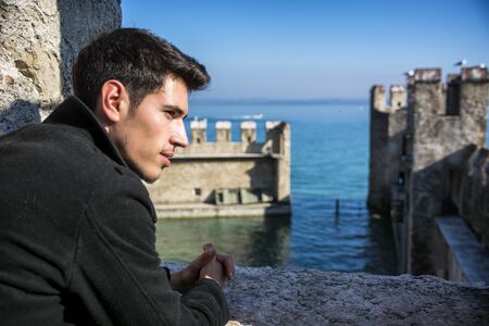 Attractive young man leaning on brick wall, in historic castle on Lake Garda in Sirmione, Italy, looking to a sideの写真素材