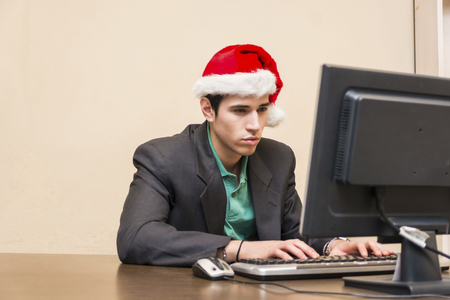 Busy young businessman sitting at his desk wearing Santa Claus red hat using computerの写真素材