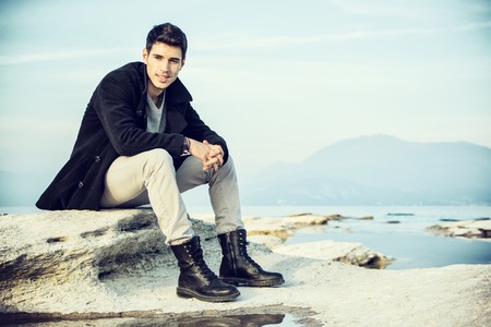 Handsome young man on a lake's shore at dusk of a sunny, peaceful day, sitting on rockの写真素材