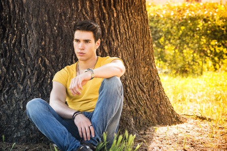 Attractive young man in park resting or relaxing against tree, in a sunny summer dayの写真素材