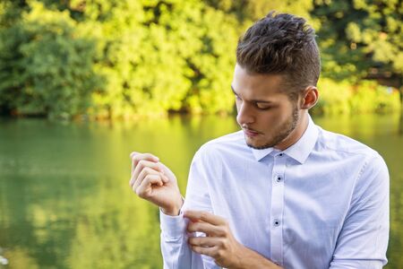Portrait of contemplative light brown haired young man wearing white shirt beside picturesque river or lake, looking at shirt cuffの写真素材