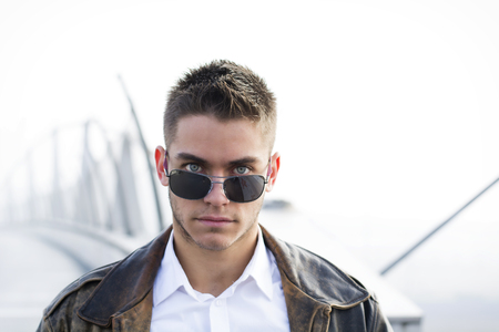 Handsome trendy young man standing on a sidewalk, wearing sunglasses and a fashionable leather jacket in a relaxed confident pose looking at camera over the glassesの写真素材