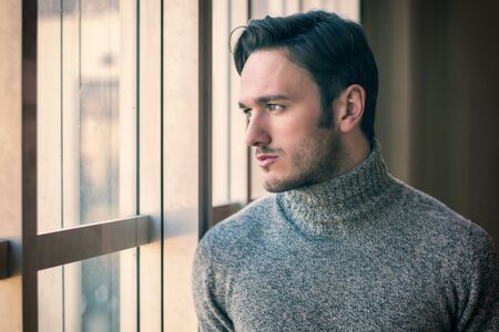 Handsome serious man standing inside modern building next to big window, wearing wool sweater,  looking outの写真素材