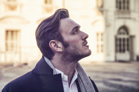 Handsome trendy man wearing dark coat standing outdoor in European city setting with elegant old historic building behind himの写真素材