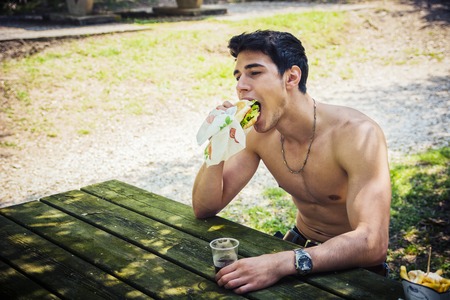 Portrait of Young Muscular Man Sitting Shirtless in Park at Picnic Table Eating Fast Food Lunch -...