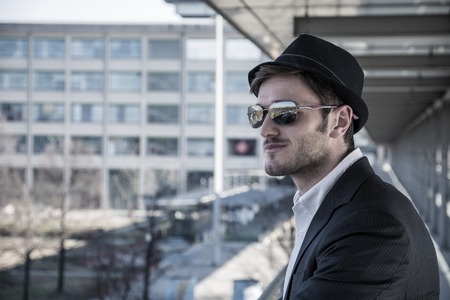 Head and Shoulders Portrait of Stylist Young Man Wearing Suit and Hat Looking to the Side Out Window While Standing on Moving Sidewalk in Buildingの写真素材