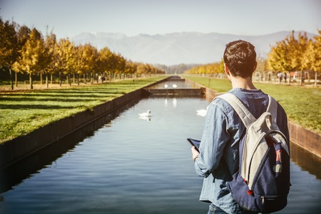 Handsome young man wearing a backpack sightseeing in a lush green autumn park, standing alongside a canal with swans holding a map or brochure, looking into the distance as though lostの写真素材