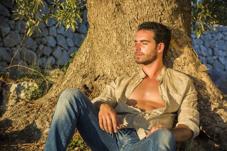 Ruggedly Handsome Man Seated at Base of Tree with Shirt Unbuttoned and Eyes Closed Relaxing in Warm Sunlightの写真素材