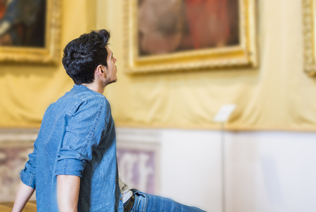 Half Body Shot of a Thoughtful Handsome Young Man, Looking At Painting Sitting on Bench Inside a Museumの写真素材