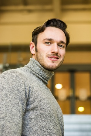 Waist Up Portrait of Attractive Young Man Wearing Sweater Standing with Hand on Chin Looking Thoughtful in Hallway of School with Students in Backgroundの写真素材