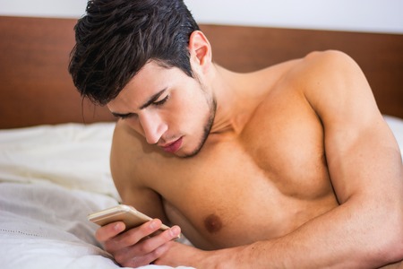Handsome young man in bed typing on cell phone, sending text message or dialing numberの写真素材
