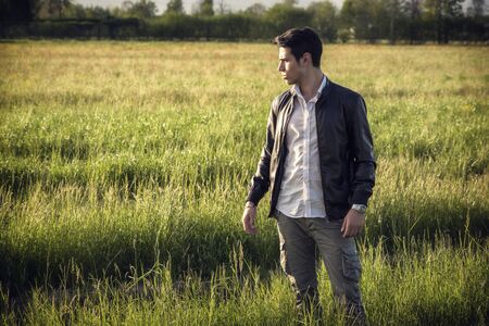 Handsome young man at countryside, in front of field or grassland, wearing white shirt and jacket, looking away in the distanceの写真素材