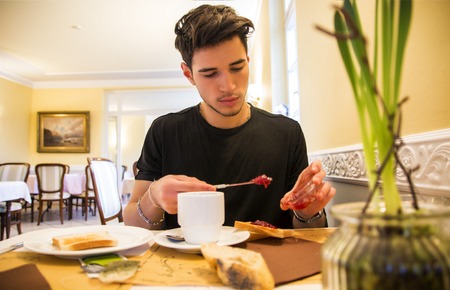 Attractive Young Man Eating Breakfast, Having a Slice of Bread with Jam and Butter and Drinking Coffeeの写真素材