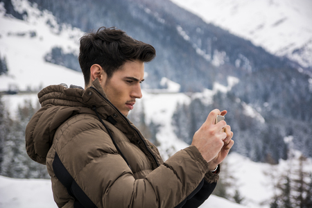 Side view on dark haired young man in outerwear taking photo of landscape in mountain with smartphoneの写真素材