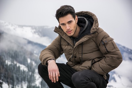 Handsome man in outerwear sitting while looking at camera. Snowy landscape on backgroundの写真素材