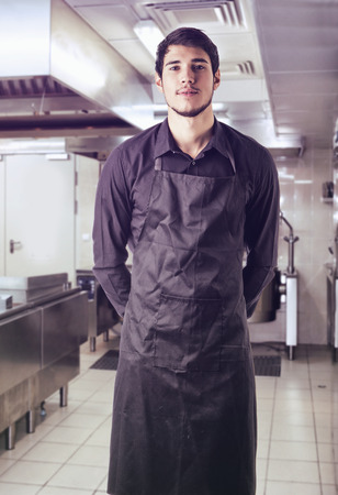 Young chef or waiter posing, wearing black apron and shirt standing in restaurant kitchenの写真素材