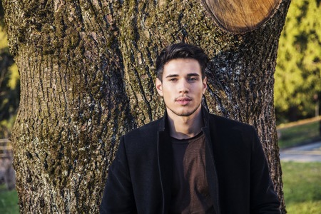 Handsome young man leaning against tree, looking at camera, in a sunny day wearing a black coatの写真素材