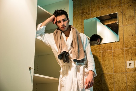 Handsome young man wearing white bathrobe, standing in spa bath next to closetの写真素材