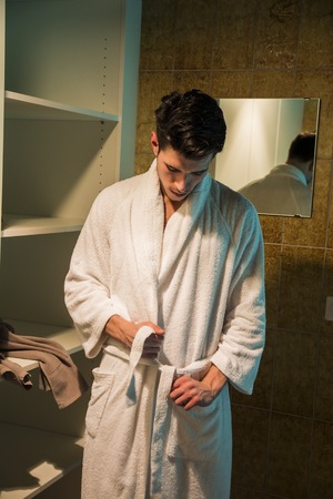 Handsome young man wearing white bathrobe, standing in spa bath next to closet
