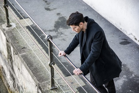 Sad brunet in black clothes looking down while standing near railing outdoors.