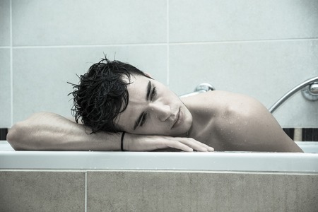 Handsome young man in bathtub at home having bath, washing body and hair with bathfoam and shampooの写真素材