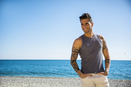 Half Body Shot of a Handsome Athletic Young Man in Trendy Attire, on a Beach in a Sunny Summer Day, Looking At Camera, Smiling against Blue Sky Background.の写真素材