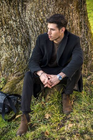Handsome young man leaning against tree, looking at camera, in a sunny day wearing a black coatの写真素材