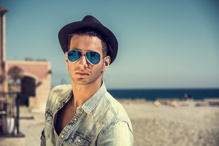 Three Quarter Shot of a Handsome Athletic Young Man in Trendy Attire, on a Beach in a Sunny Summer Day, Looking At Camera wearing sunglasses and hat, against Blue Sky Background.の写真素材