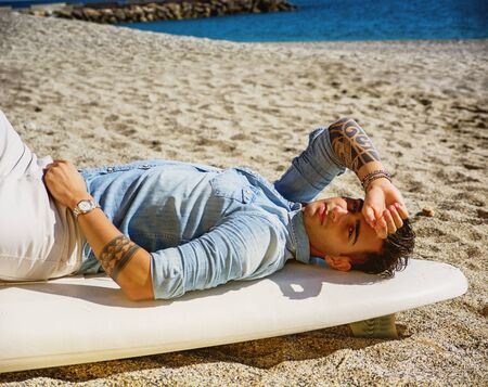 Stylish handsome young man laying on surfboard on beach in sunlightの写真素材