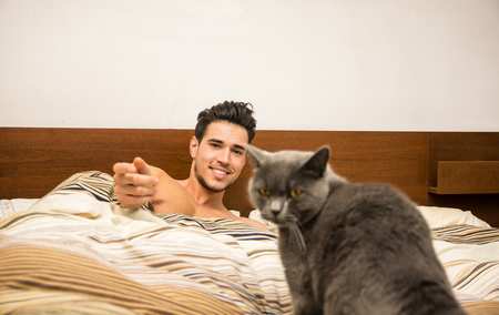 Handsome Young Animal-Lover Man on a Bed, Hugging and Cuddling his Gray Domestic Cat Pet.の写真素材