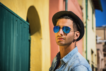 Portrait of dark haired young man in blue mirrored sunglasses and black hat. Cityscape on background, with colorful houses in Italyの写真素材