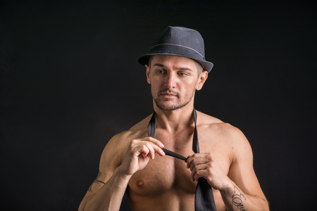 Handsome shirtless muscular man standing on dark background, wearing jeans, fedora hat and neck-tie on naked torsoの写真素材