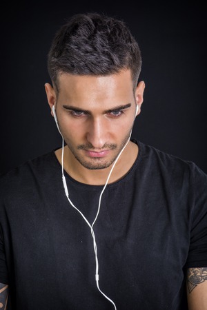 Attractive young man with earphones listening to music, looking down, on dark backgroundの写真素材