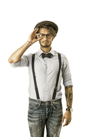 Portrait of brunette young man in glasses, hat, bow-tie, suspenders and shirt looking at camera. Studio shot, isolated on whiteの写真素材