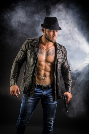 Handsome man wearing leather jacket on naked muscular torso, holding hand gun, on dark smoky background, looking to a side. Wearing fedora hatの写真素材