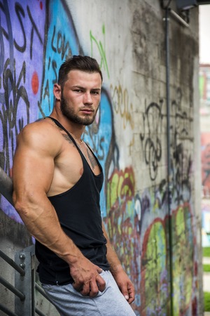 Handsome Muscular Hunk Man Outdoor in City Setting. Showing Healthy Body While Looking at Camera, Leaning against Graffiti Wallの写真素材